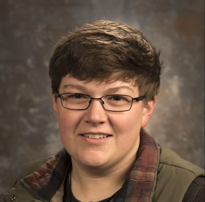 A white woman with short straight brown hair wearing a work vest with a flannel collar, brown shirt, and square glasses.