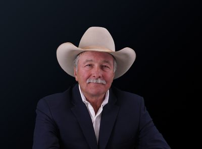 A white man in a well-fitted suit and a felt tan cowboy hat. He has gray hair and a short mustache.