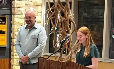A young thin white woman with shoulder length straight blonde hair stands at a podium giving a speech. Next to the podium, a tall, broad, bald white man with a short beard wearing a sweater and glasses is standing by, looking proud.