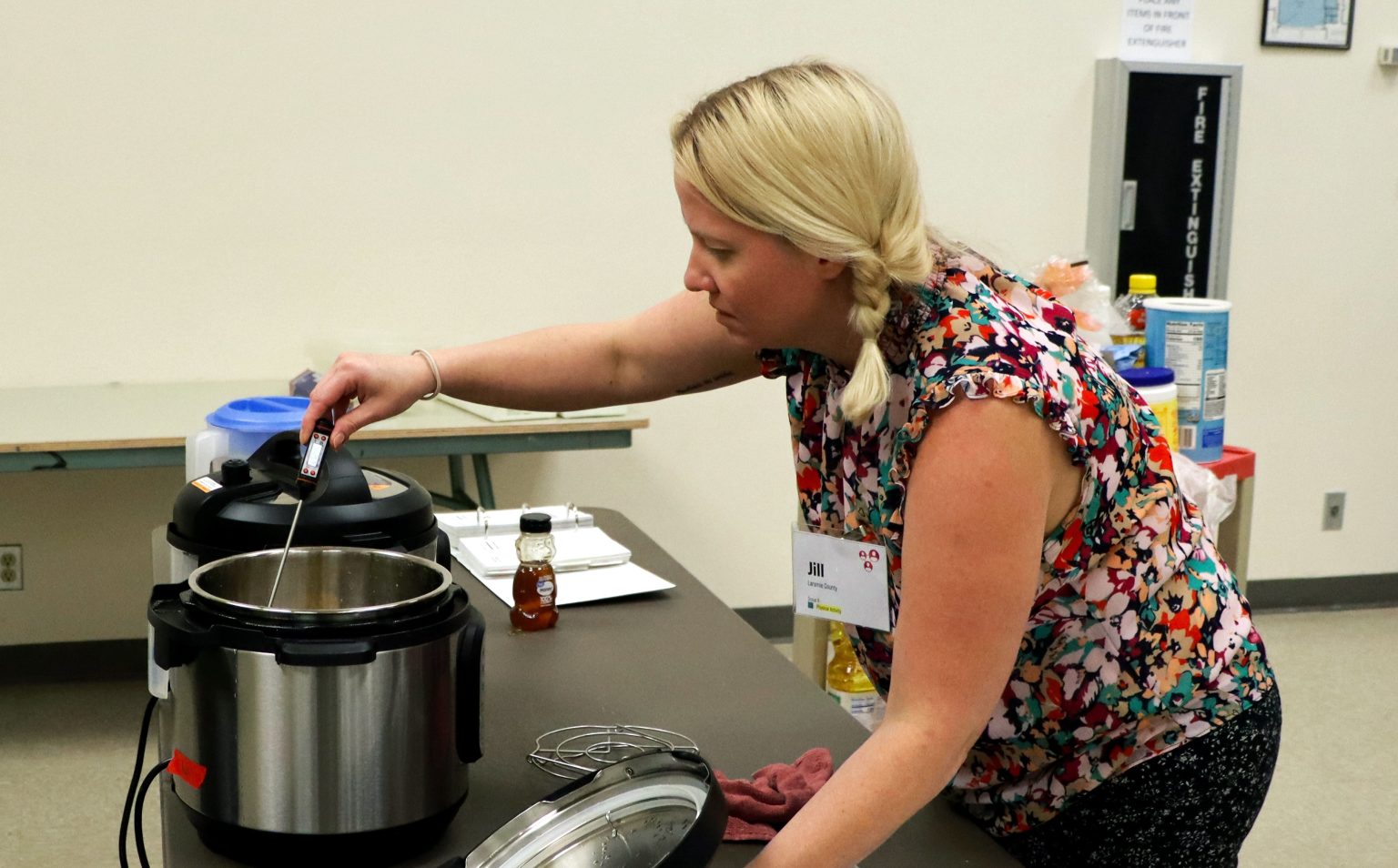 blond woman wearing floral shirt and name tag that says Jill holds an Instant Pot lid in one hand and sticks a food thermometer into the electric pressure cooker with the other hand