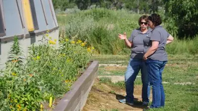 two women wearing dark gray polo shirts and jeans stand beside a flower bed with yellow flowers alongside a geodome greenhouse