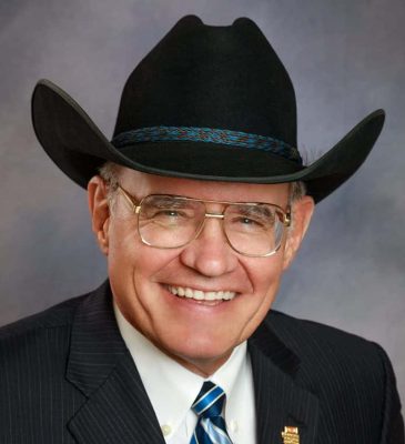 An older white man in a black suit and black cowboy hat with a striped blue tie. He has rounded features, is smiling jovially, and is wearing square glasses.