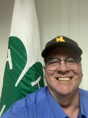 A stocky white man with round glasses smiling. He is wearing a brown and gold hat with Steamboat (the bucking horse) on it and is standing in front of a 4-H flag. 