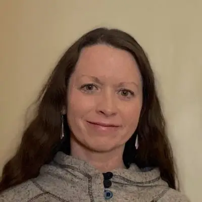 An image of a white woman with long fine wavy brown hair. She is wearing earrings and a fancy gray sweater.
