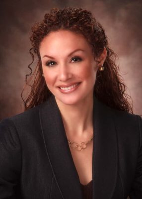 An image of a white woman with long curly brown hair. She is wearing makeup, a black suit jacket, and a small gold necklace and earrings. 