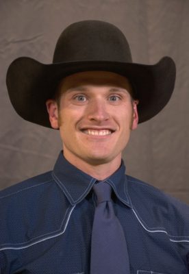 An image of a young white man wearing a large black cowboy hat and a dark blue dress shirt and tie.