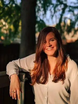 A young white woman with long blondeish brown hair in an outdoor setting. She is wearing a white cardigan.