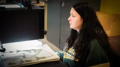 A large sheet of paper with a pressed plant on it is placed underneath a large black machine, which shines light at it, by a young woman with long black hair.