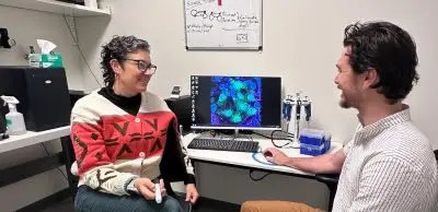 Two people talking in a small office/lab space. On the left, a middle aged woman with short curly black hair and glasses wearing a cardigan with bold red patterns. On the right, a young man with short wavy brown hair and short facial hair wearing a white button up shirt with vertical stripes and brown pants.