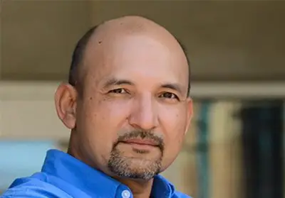 A middle-aged man with light brown skin, a black goatee, and a shaved head. He is wearing a blue polo t-shirt.