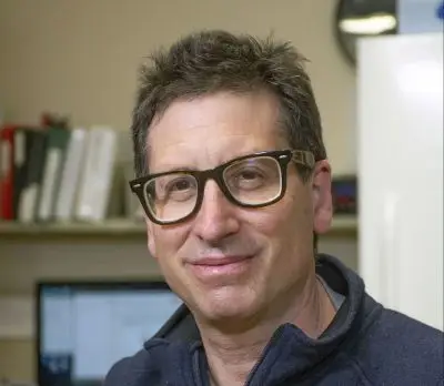 White man with short brown-gray hair and thick square glasses wearing a gray vest and long-sleeve shirt.