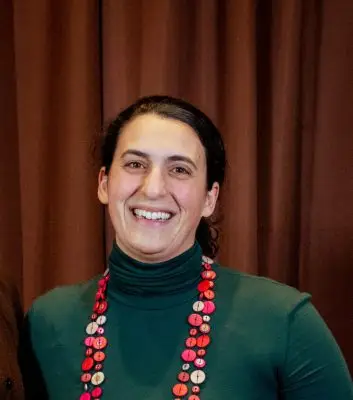 A woman with light skin and curly black hair pulled back. She is wearing a green turtleneck and a long red necklace.