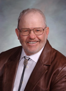 A middle aged white man with round wire rim glasses wearing a bolo tie and a brown leather suit jacket. He has some stubble and is mostly bald.