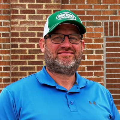 A white man wearing a blue polo shirt and green baseball cap with the words "Kings Rope - Sheridan, Wyoming" on it. He has square glasses and a beard and is middle aged.