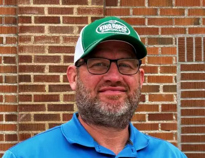 A white man wearing a blue polo shirt and green baseball cap with the words "Kings Rope - Sheridan, Wyoming" on it. He has square glasses and a beard and is middle aged.
