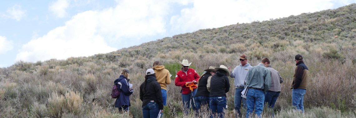 instructor wearing baseball cap, sweatshirt, and jeans addresses a group of students standing on a hill covered in sagebrush with a mostly overcast sky above