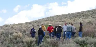 instructor wearing baseball cap, sweatshirt, and jeans addresses a group of students standing on a hill covered in sagebrush with a mostly overcast sky above