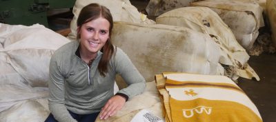 A young white woman sitting down in several burlap bags. On one of them there is a white, brown, and gold blanket marked "UW".