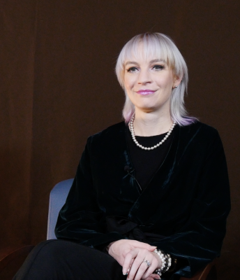 A woman with very light skin and platinum blonde shoulder length hair that is dyed a purplish pink color at the tips. She is wearing a bright purplish lipstick, a pearl necklace, and a black long sleeve shirt.