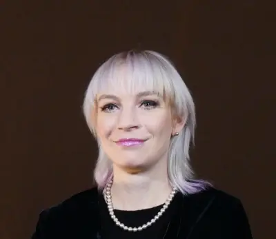A woman with very light skin and platinum blonde shoulder length hair that is dyed a purplish pink color at the tips. She is wearing a bright purplish lipstick, a pearl necklace, and a black long sleeve shirt.