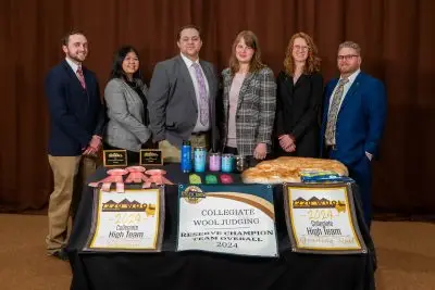 five young people and their professor, all in formal garb, stand behind a table displaying plaques, award ribbons, waterbottles, and a sheep fleece