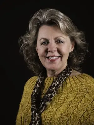 A woman with light skin, wavy silver chin-length hair, wearing a yellow sweater and scarf.