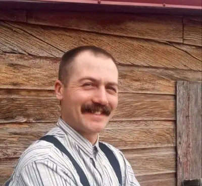 A young man with light skin, short cropped hair, and a mustache wearing a button up striped shirt and suspenders. He is standing next to a wooden building.