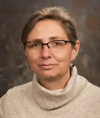 portrait of smiling woman with brown hair and glasses wearing beige sweater and dangly earrings