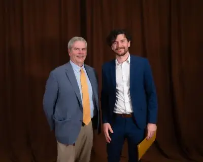 A man with short gray hair wearing a suit and a young man with wavy short brown hair and a short beard/mustache wearing a blue suit.