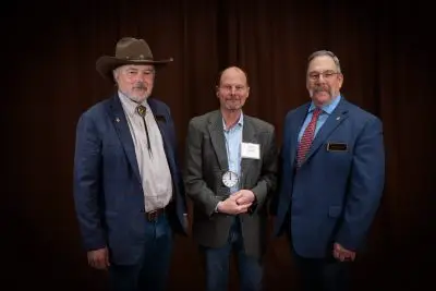 Three white men in formal wear. On the left, a man with a white beard wearing a cowboy hat and bowler. In the middle, a shorter man holding a plaque with cropped hair and a goatee. On the right, a man with short gray hair and a handlebar mustache wearing wire rim glasses.