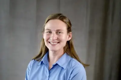 A woman with shoulder length dirty blonde hair and light skin wearing a light blue collared shirt.