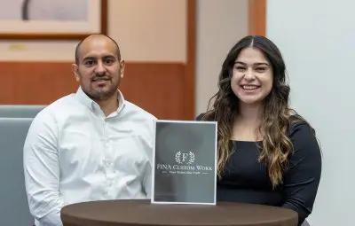 A man and a woman with light brown skin standing behind a black plaque that reads "Fina Custom Work: Your vision, our craft". The woman has long wavy black hair with the tips dyed brown and the man has very short hair and stubble.