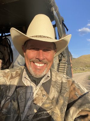 An older man with a goatee and tan skin wearing a tall white cowboy hat and camo.