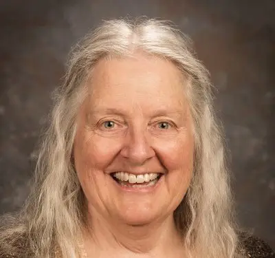 An older woman with light skin and shoulder length gray hair with slight waves. She is wearing a shiny gold shirt and a brown cardigan.