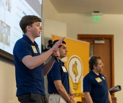Three men giving a presentation. On the left, a young white man with short wavy brown hair. In the middle, a young man with tan skin and short very black straight hair. On the right, an older man with gray wavy hair, a beard, and glasses.