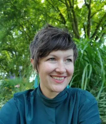 A woman with light skin and short straight dark brown hair. She is wearing large teal and white earrings and a turquoise long sleeve shirt. The background is plants.