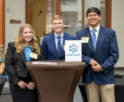 Three young people in formal wear standing behind a white and blue plaque that reads "Power Theory". On the left, a woman with light skin and wavy blonde hair slightly beyond her shoulders. In the middle, a man with light skin and short blonde hair. On the right, a tall man with light brown skin, glasses, and short black hair.