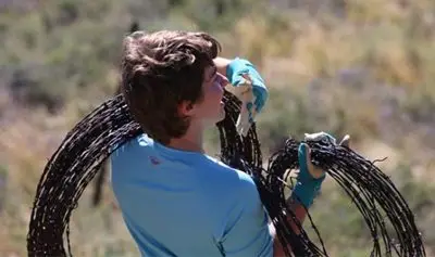 A young person with short straight brown hair and light skin holding two coils of barbed wire. She has one over her shoulder and another in a gloved hand.