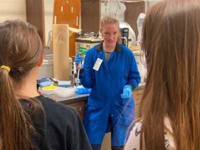 A white woman in a blue jumpsuit and plastic gloves holding up a syringe. In the background, there is lab equipment. Two girls are looking at the woman in blue.