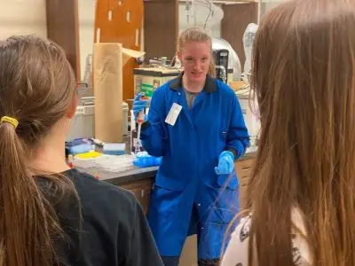 A white woman in a blue jumpsuit and plastic gloves holding up a syringe. In the background, there is lab equipment. Two girls are looking at the woman in blue.