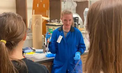 A white woman in a blue jumpsuit and plastic gloves holding up a syringe. In the background, there is lab equipment. Two girls are looking at the woman in blue.