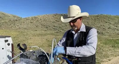Finley using plastic hoses to add fluid to two metal canisters out on a dirt road.