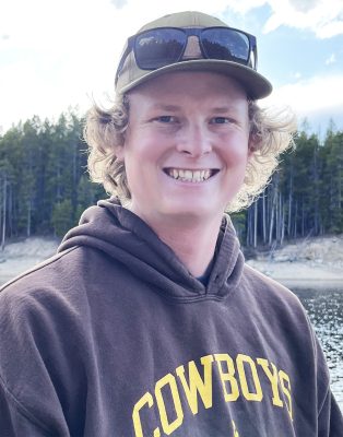 A young man with light skin and short curly blonde hair wearing a Wyoming Cowboys hoodie and a baseball cap with sunglasses on top. He is standing in front of a lake. 
