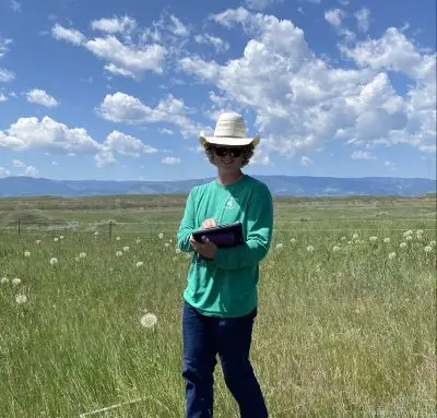 A man wearing a cowboy hat and jeans standing in a grassy field with a clipboard on a sunny day.
