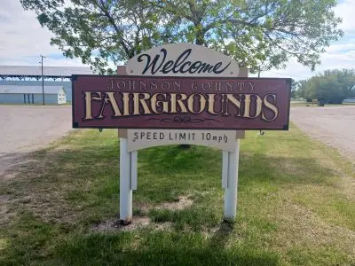 A sign that reads "Welcome - Johnson County Fairgrounds - Speed Limit 10mph" in fancy tan, brown, and gold lettering. The sign is in a park-like area with trees, roads, buildings.