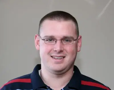 A man with cropped blonde hair, glasses, and light skin wearing a striped collared shirt.