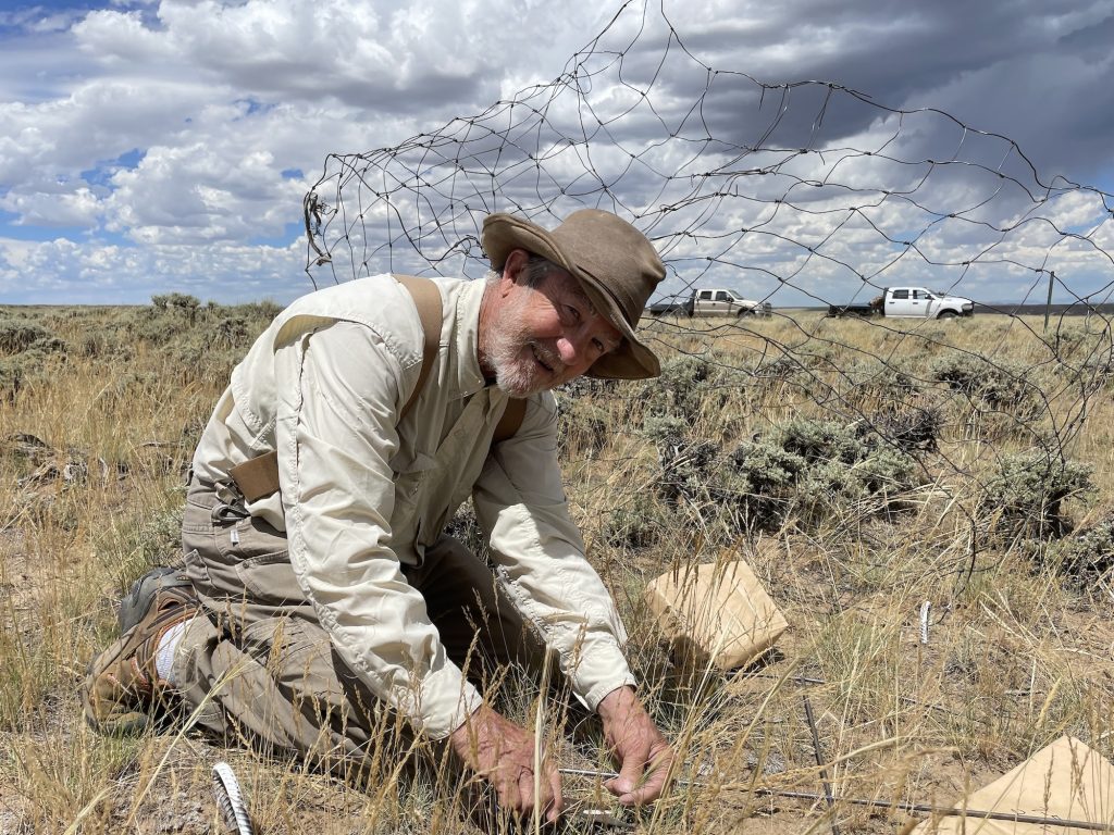 smiling older man wearing a brown hat, long-sleeved tan shirt, suspenders, and brown pants kneels on the ground beside a tangled piece of wire fencing, clipping vegetation samples within a circular metal hoop
