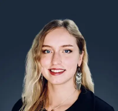 A woman with long wavy blonde hair and light skin wearing red lipstick, earrings, a necklace, and a black shirt.