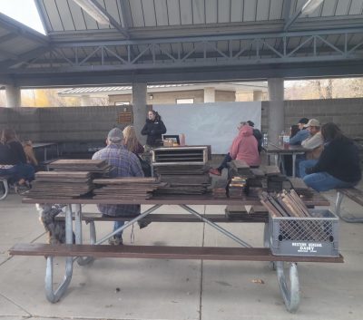A picnic shelter in which ten people listen to a young woman wearing a black hoodie. In the foreground, one picnic table is stacked with short dark brown planks. 