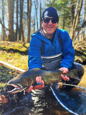 A man wearing athletic clothes and sunglasses holding a fish that is about a foot and a half long. 